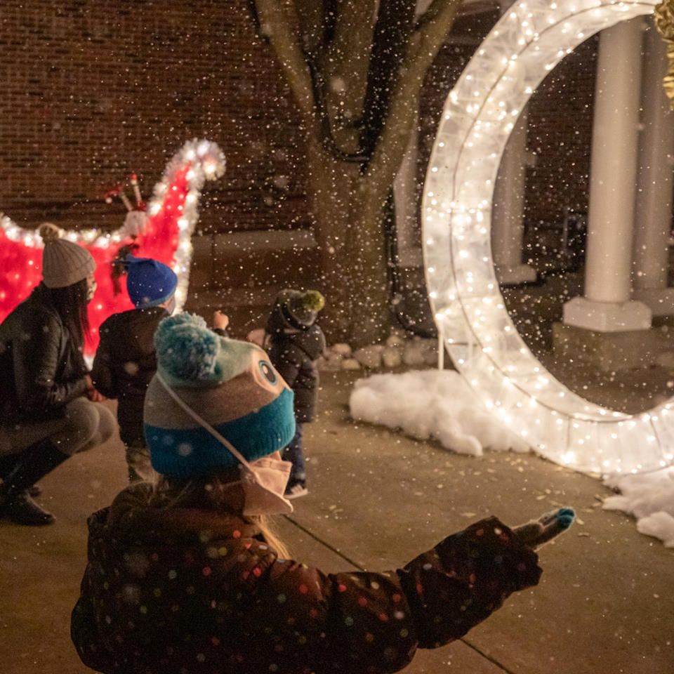 Snow falls at outdoor light display. Boy dressed in winter gear reaches for snowflake.