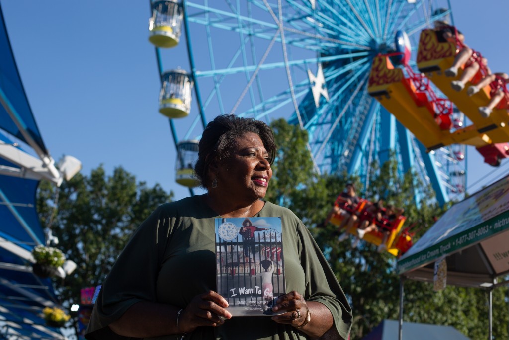 Denise Montgomery stands in front of a Ferris Wheel at Fair Park in Dallas, holding a copy of the book she wrote, titled I Want To Go. 