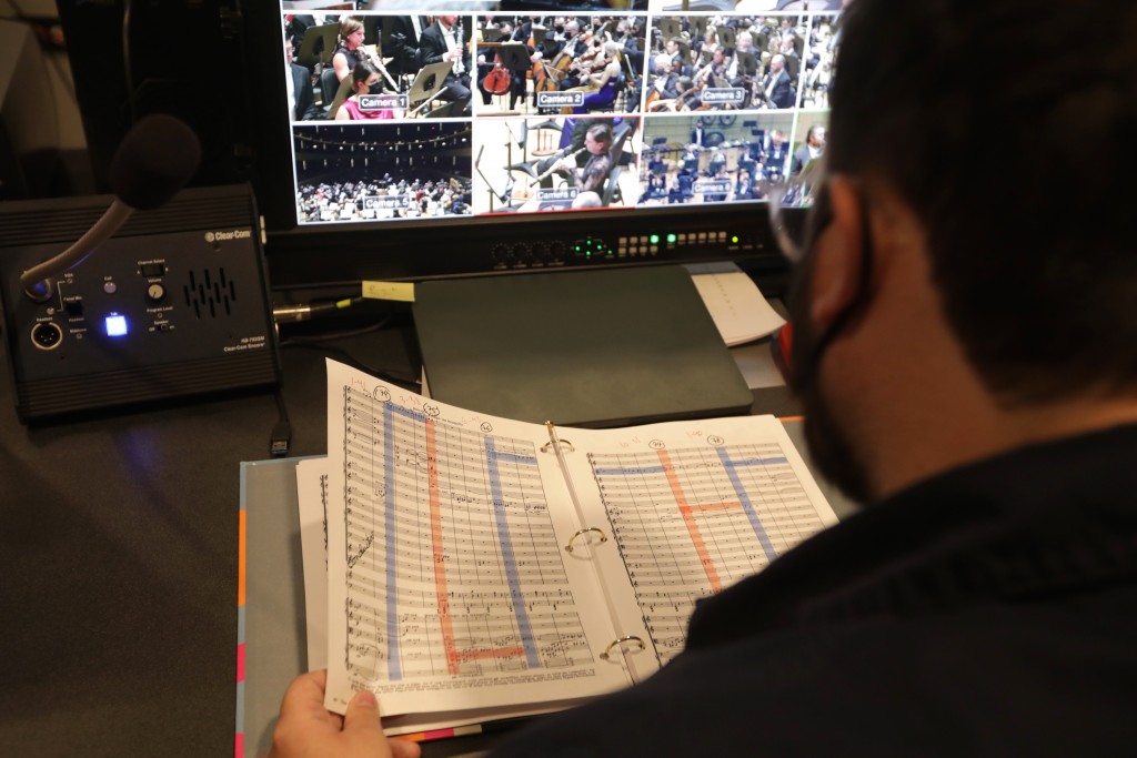 Andrew Alden is directing videos by looking at a score sheet in a binder and the camera angles displayed on a video screen.